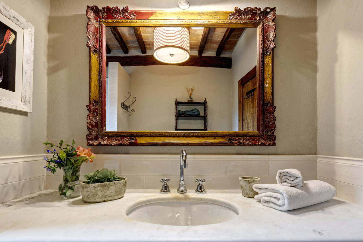 Elegant bathroom sink area featuring a vintage-style mirror with ornate frame, modern faucet, and neatly arranged white towels. Decorative plants and flowers add a touch of freshness to the marble countertop, enhancing the overall aesthetic. Natural light illuminates the space, showcasing wooden beams and a cozy atmosphere. | Only on SopranoVillas