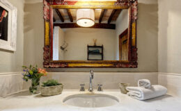 Elegant bathroom sink area featuring a vintage-style mirror with ornate frame, modern faucet, and neatly arranged white towels. Decorative plants and flowers add a touch of freshness to the marble countertop, enhancing the overall aesthetic. Natural light illuminates the space, showcasing wooden beams and a cozy atmosphere.