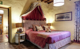 Cozy bedroom featuring a king-sized bed with a striped headboard, adorned with decorative pillows and a rich burgundy bedspread. The room includes wooden accents, a bedside table with books, and ambient lighting from lamps. Natural textures and warm tones create an inviting atmosphere, complemented by patterned curtains and a vintage wall art piece.