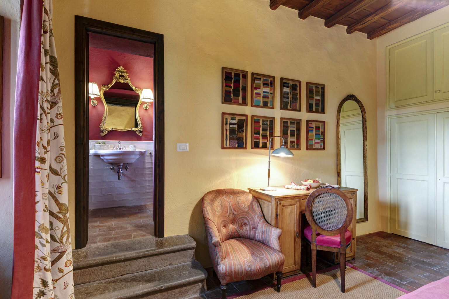 Cozy interior of a stylish room featuring a decorative chair, wooden desk with a lamp, and colorful wall art. A mirror and a glimpse of a bathroom can be seen through the doorway, highlighting rustic charm and warm tones. Ideal for showcasing home décor and interior design aesthetics. | Only on SopranoVillas