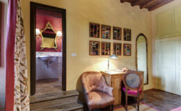 Cozy interior of a stylish room featuring a decorative chair, wooden desk with a lamp, and colorful wall art. A mirror and a glimpse of a bathroom can be seen through the doorway, highlighting rustic charm and warm tones. Ideal for showcasing home décor and interior design aesthetics.