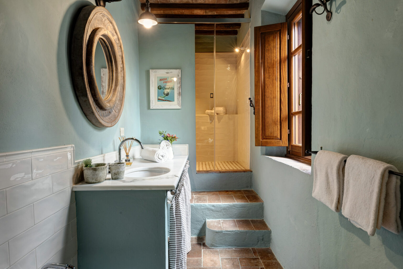 Modern bathroom interior featuring a stylish wooden mirror, a marble sink with decorative plants, and a glass shower. The walls are painted in a soft blue, complemented by warm wooden accents and terracotta flooring, creating a cozy and inviting atmosphere. | Only on SopranoVillas