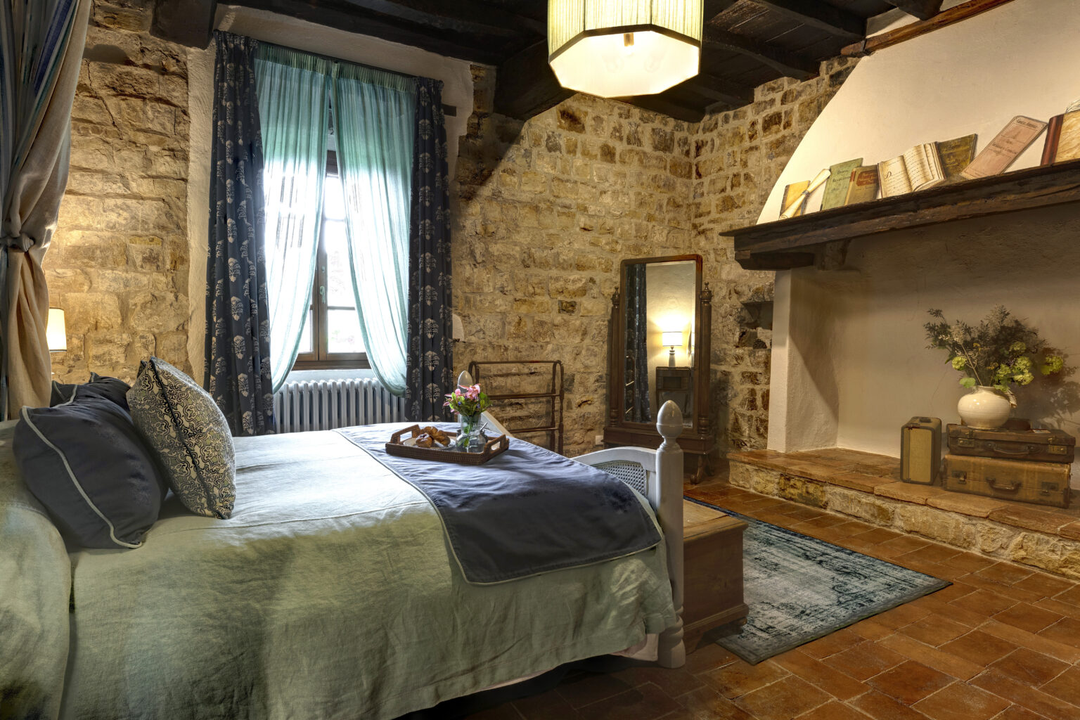 Cozy bedroom featuring rustic stone walls, a comfortable bed with decorative pillows, and large windows dressed with sheer curtains. The room includes a vintage mirror, a warm-toned wooden shelf with books, and a charming arrangement of flowers, creating a serene and inviting atmosphere. | Only on SopranoVillas
