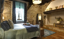 Cozy bedroom featuring rustic stone walls, a comfortable bed with decorative pillows, and large windows dressed with sheer curtains. The room includes a vintage mirror, a warm-toned wooden shelf with books, and a charming arrangement of flowers, creating a serene and inviting atmosphere.