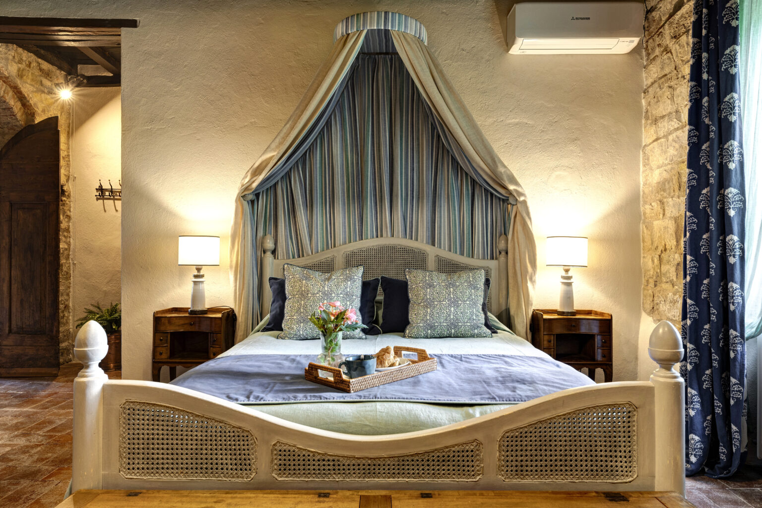 Cozy bedroom featuring a stylish canopy bed with decorative pillows, a bedside table with a lamp, and a tray with flowers and pastries. The room has a warm ambiance with stone walls and patterned curtains, ideal for relaxation and comfort. | Only on SopranoVillas