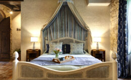 Cozy bedroom featuring a stylish canopy bed with decorative pillows, a bedside table with a lamp, and a tray with flowers and pastries. The room has a warm ambiance with stone walls and patterned curtains, ideal for relaxation and comfort.