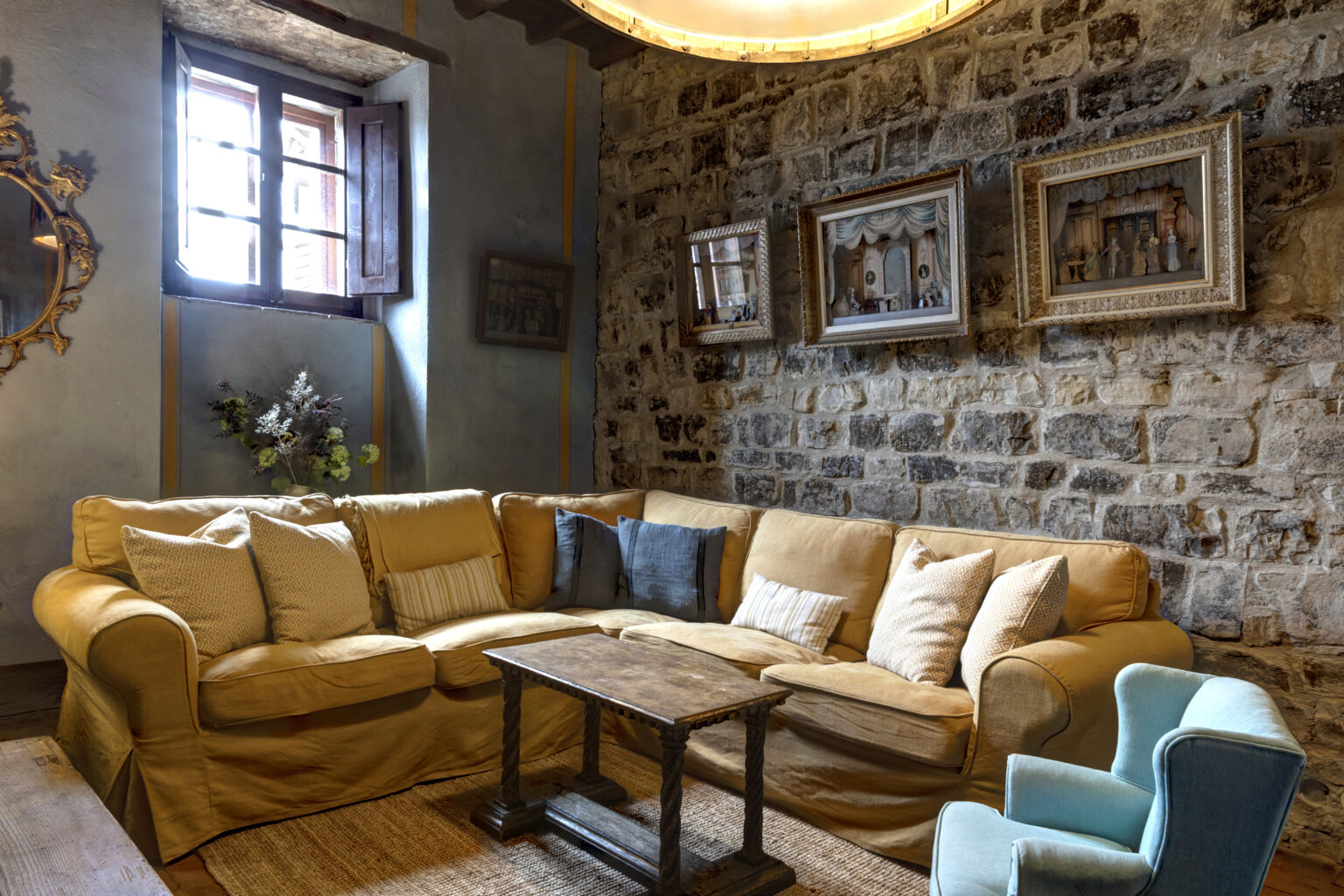 Cozy living room featuring a yellow sofa with decorative pillows, a rustic wooden coffee table, and a light blue armchair. The stone wall background is adorned with framed artwork, and a window allows natural light to illuminate the space, creating a warm and inviting atmosphere. | Only on SopranoVillas