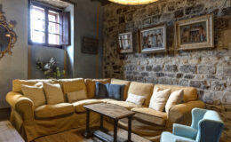 Cozy living room featuring a yellow sofa with decorative pillows, a rustic wooden coffee table, and a light blue armchair. The stone wall background is adorned with framed artwork, and a window allows natural light to illuminate the space, creating a warm and inviting atmosphere.