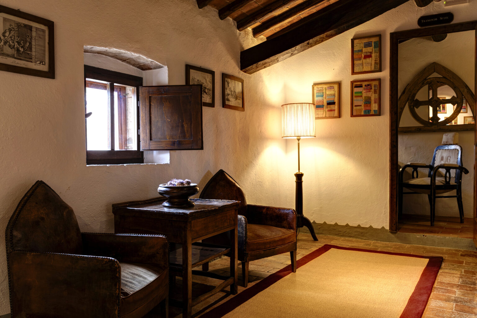 Cozy interior of a rustic room featuring two vintage chairs, a wooden table, and a softly lit lamp, complemented by artwork on the walls and an open window allowing natural light. | Only on SopranoVillas
