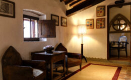 Cozy interior of a rustic room featuring two vintage chairs, a wooden table, and a softly lit lamp, complemented by artwork on the walls and an open window allowing natural light.
