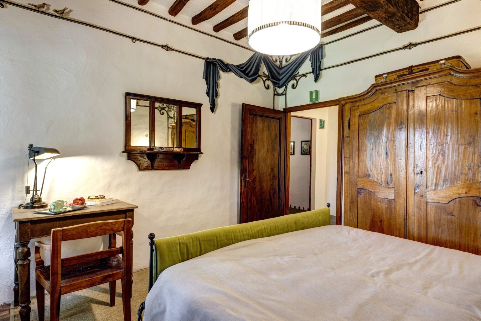 Cozy bedroom featuring a green upholstered bed, wooden furniture including a desk and chair, a vintage mirror, and rustic decor elements. The room has exposed wooden beams and warm lighting, creating a welcoming atmosphere. | Only on SopranoVillas