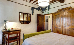 Cozy bedroom featuring a green upholstered bed, wooden furniture including a desk and chair, a vintage mirror, and rustic decor elements. The room has exposed wooden beams and warm lighting, creating a welcoming atmosphere.