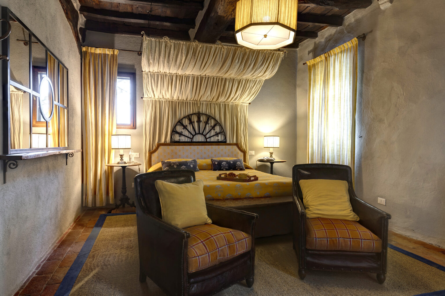 Cozy bedroom interior featuring a queen-sized bed with yellow bedding, two comfortable armchairs, and large windows adorned with sheer curtains. The room is illuminated by soft lighting from bedside lamps and a unique ceiling light, creating a warm and inviting atmosphere. Rustic wooden beams and textured walls enhance the charming decor. | Only on SopranoVillas