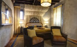 Cozy bedroom interior featuring a queen-sized bed with yellow bedding, two comfortable armchairs, and large windows adorned with sheer curtains. The room is illuminated by soft lighting from bedside lamps and a unique ceiling light, creating a warm and inviting atmosphere. Rustic wooden beams and textured walls enhance the charming decor.