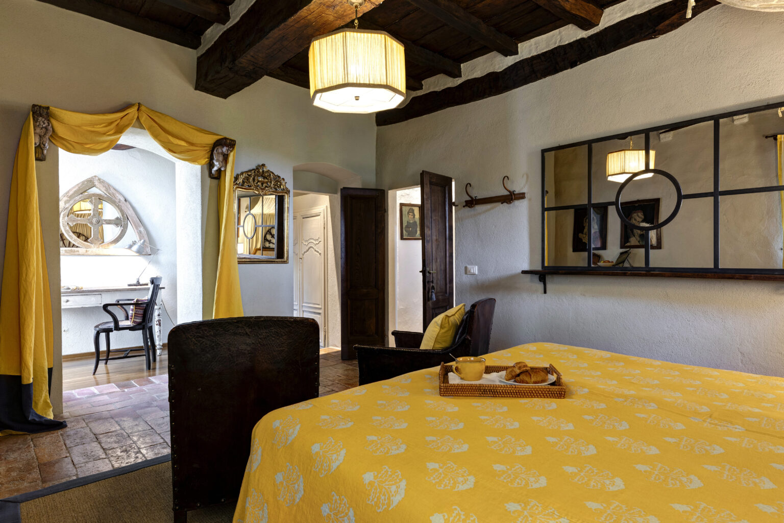 Cozy bedroom featuring a yellow floral bedspread, a comfortable chair, and a breakfast tray with pastries. The room includes rustic wooden beams, a decorative mirror, and a workspace with a stylish chair, creating a warm and inviting atmosphere. | Only on SopranoVillas