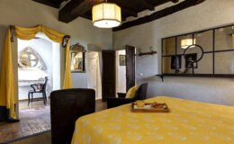 Cozy bedroom featuring a yellow floral bedspread, a comfortable chair, and a breakfast tray with pastries. The room includes rustic wooden beams, a decorative mirror, and a workspace with a stylish chair, creating a warm and inviting atmosphere.
