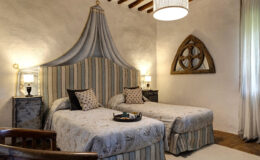 Cozy bedroom featuring two twin beds with elegant patterned bedding, a decorative headboard draped with fabric, rustic wooden accents, and soft lighting, creating a warm and inviting atmosphere.