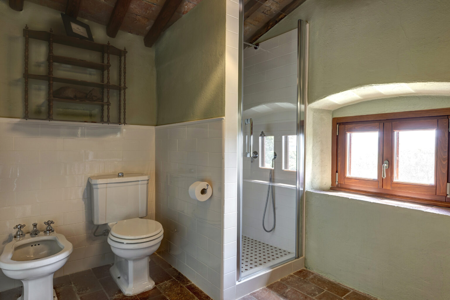 Modern bathroom featuring a white toilet and bidet, a glass shower enclosure, and a rustic wooden shelf. Natural light streams in through a window, highlighting the elegant tile work and earthy color palette. Ideal for showcasing contemporary home design. | Only on SopranoVillas