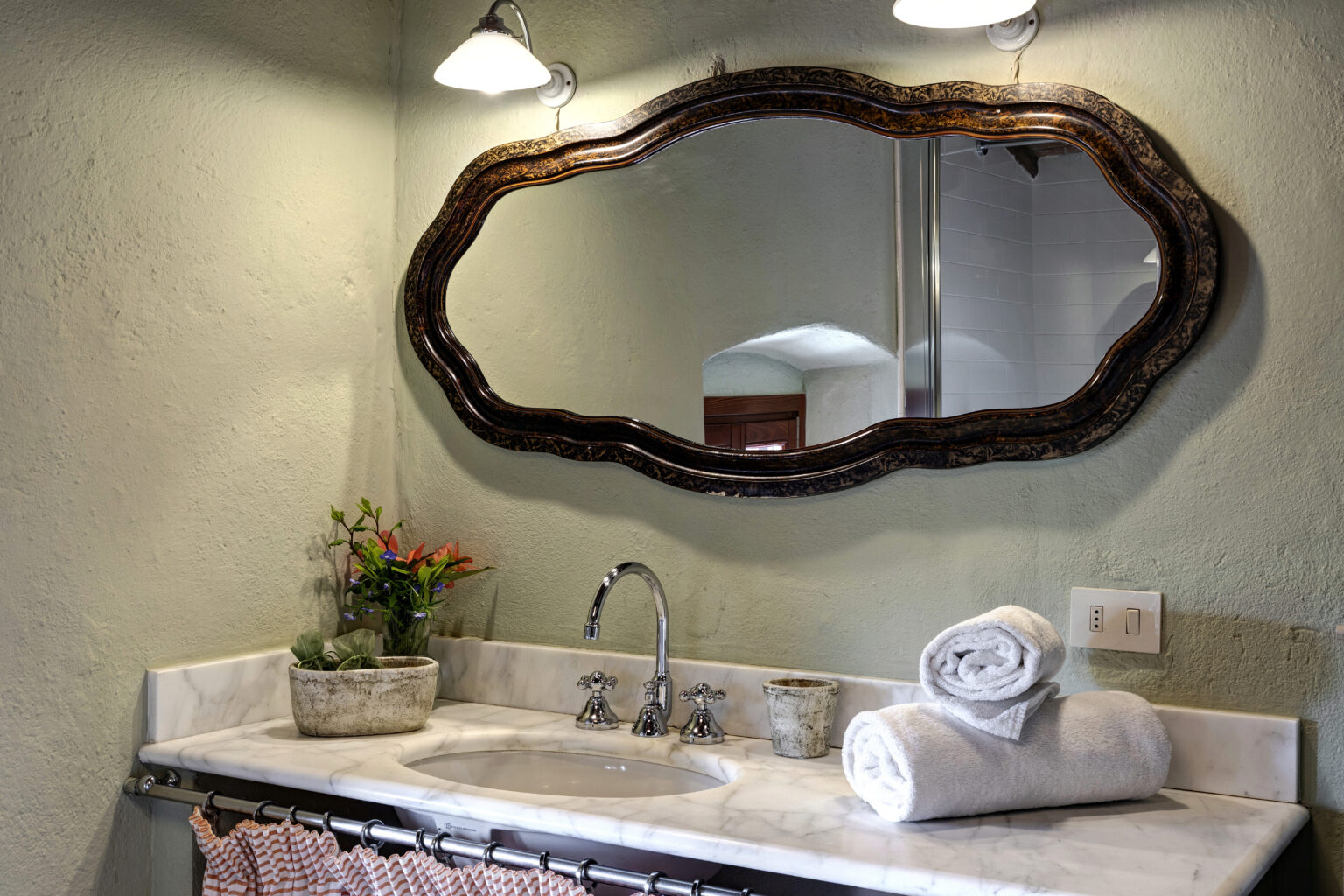 Elegant bathroom vanity featuring a vintage oval mirror, chrome faucet, and neatly rolled white towels on a marble countertop. Decorative potted plants add a touch of greenery, enhancing the serene atmosphere. Soft lighting illuminates the space, creating a cozy ambiance. | Only on SopranoVillas