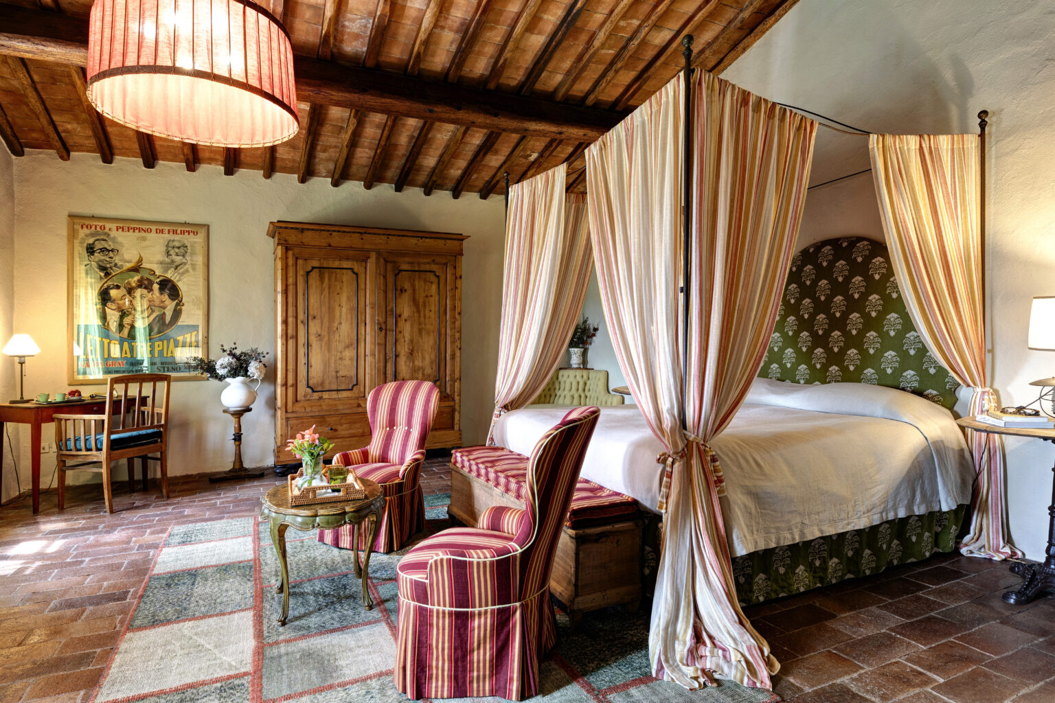 Cozy bedroom featuring a four-poster bed with striped drapes, vintage wooden furniture, and a warm color palette. The room includes a seating area with striped chairs, a coffee table, and a decorative rug, complemented by rustic wooden cabinetry and an antique lamp, creating a charming and inviting atmosphere. | Only on SopranoVillas