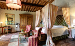 Cozy bedroom featuring a four-poster bed with striped drapes, vintage wooden furniture, and a warm color palette. The room includes a seating area with striped chairs, a coffee table, and a decorative rug, complemented by rustic wooden cabinetry and an antique lamp, creating a charming and inviting atmosphere.