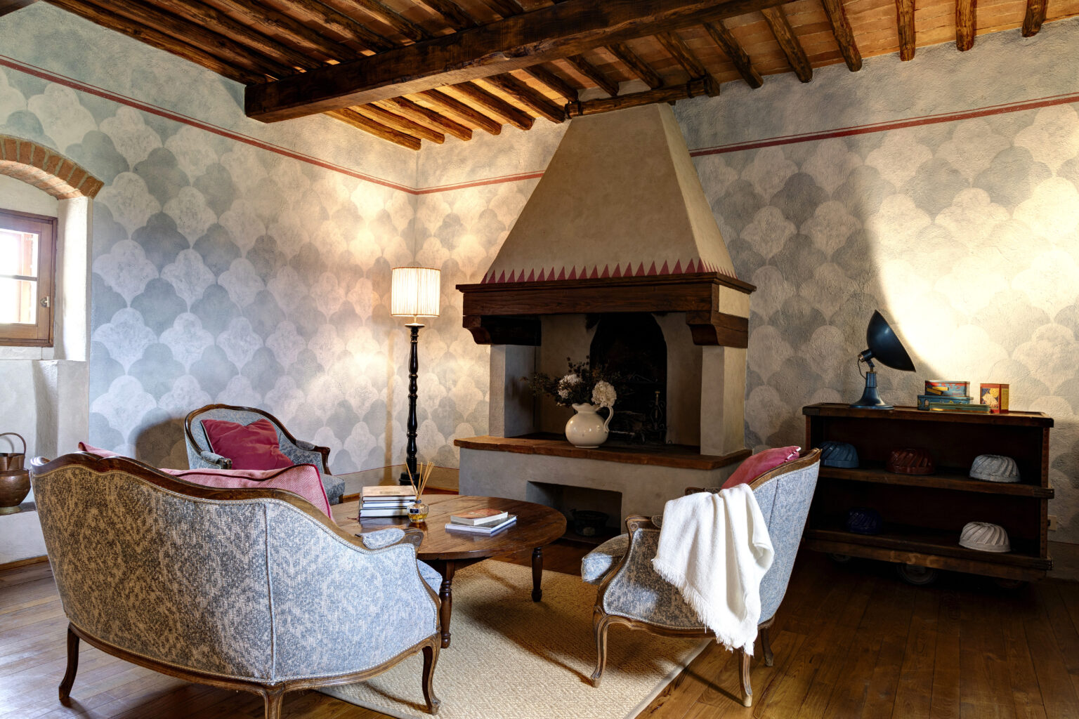 Cozy living room featuring vintage-style armchairs, a wooden coffee table, and a decorative fireplace. The walls are adorned with elegant patterned wallpaper, complemented by a stylish floor lamp and a unique blue desk lamp on a wooden shelf. Natural light enhances the warm atmosphere of the space. | Only on SopranoVillas