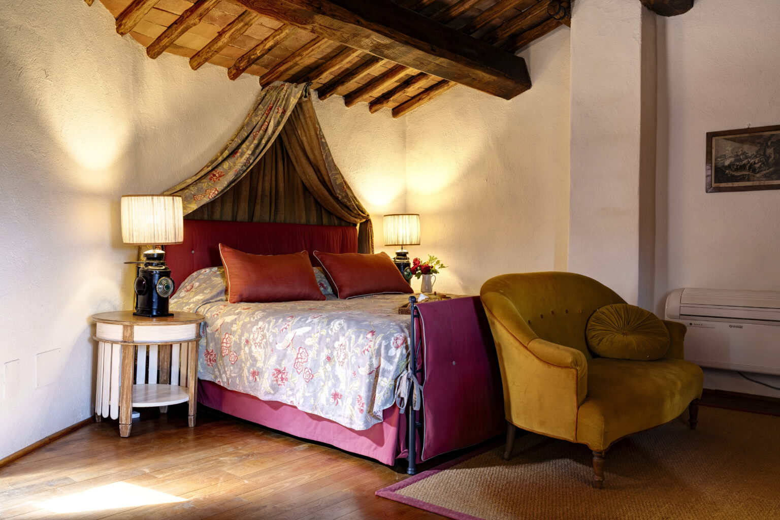 Cozy bedroom interior featuring a floral bedspread, decorative pillows, and a vintage lamp. The room has exposed wooden beams, a comfortable yellow armchair, and a small side table, creating a warm and inviting atmosphere. Ideal for relaxation and comfort. | Only on SopranoVillas