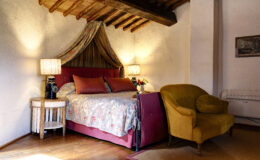 Cozy bedroom interior featuring a floral bedspread, decorative pillows, and a vintage lamp. The room has exposed wooden beams, a comfortable yellow armchair, and a small side table, creating a warm and inviting atmosphere. Ideal for relaxation and comfort.