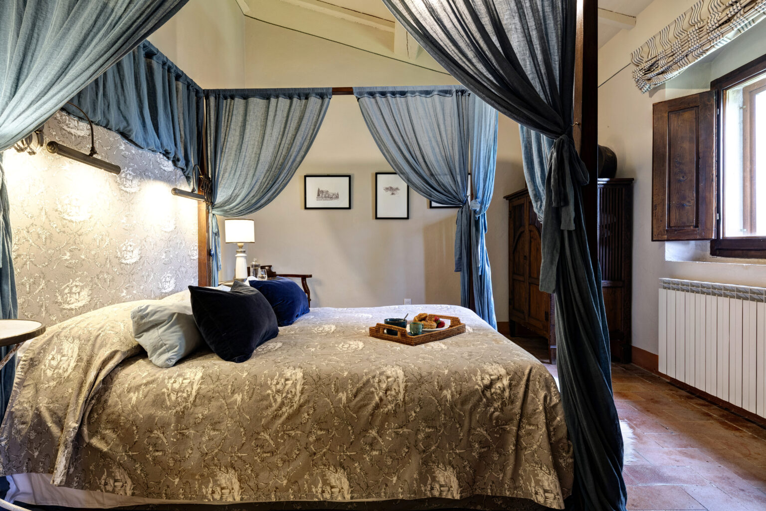 Cozy bedroom featuring a four-poster bed with blue drapes, elegant bedding, and a decorative tray with snacks. The room includes soft lighting, framed artwork, and wooden furniture, creating a warm and inviting atmosphere. Natural light streams through the window, enhancing the serene ambiance. | Only on SopranoVillas