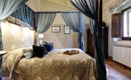 Cozy bedroom featuring a four-poster bed with blue drapes, elegant bedding, and a decorative tray with snacks. The room includes soft lighting, framed artwork, and wooden furniture, creating a warm and inviting atmosphere. Natural light streams through the window, enhancing the serene ambiance.