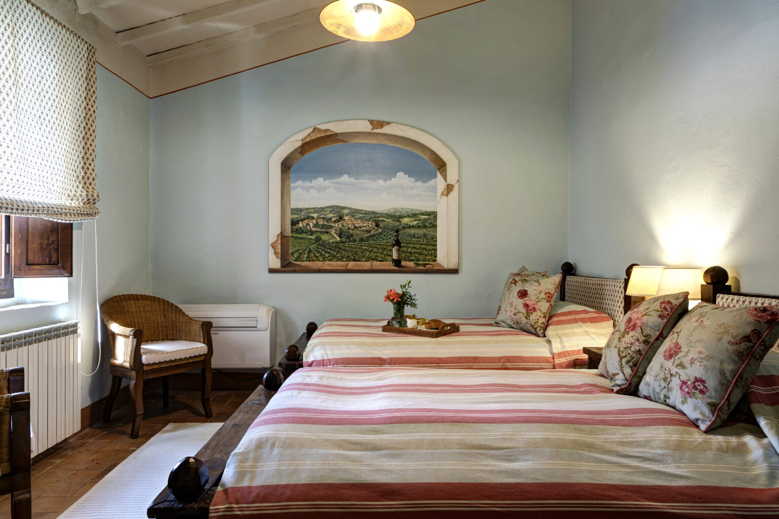 Cozy guest room featuring two striped beds with floral pillows, a wicker chair, and a scenic mural of a countryside landscape. The room includes a small table with refreshments and a warm, inviting atmosphere, perfect for relaxation. | Only on SopranoVillas