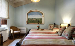 Cozy guest room featuring two striped beds with floral pillows, a wicker chair, and a scenic mural of a countryside landscape. The room includes a small table with refreshments and a warm, inviting atmosphere, perfect for relaxation.
