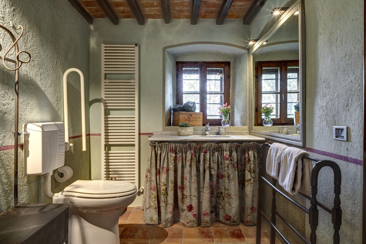 Cozy bathroom featuring a white toilet, floral sink skirt, and rustic decor. Natural light streams through wooden windows, highlighting the green walls and terracotta flooring. A heated towel rack and decorative flowers add charm. Ideal for a relaxing retreat. | Only on SopranoVillas