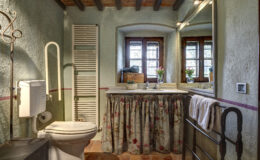 Cozy bathroom featuring a white toilet, floral sink skirt, and rustic decor. Natural light streams through wooden windows, highlighting the green walls and terracotta flooring. A heated towel rack and decorative flowers add charm. Ideal for a relaxing retreat.