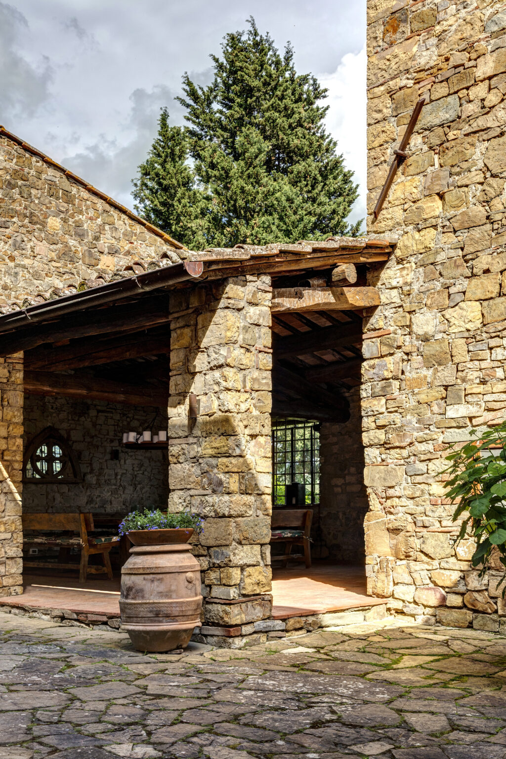 Rustic stone building with a wooden roof and an outdoor seating area, featuring a clay pot with flowers in the foreground and a tall tree in the background, set against a cloudy sky. | Only on SopranoVillas