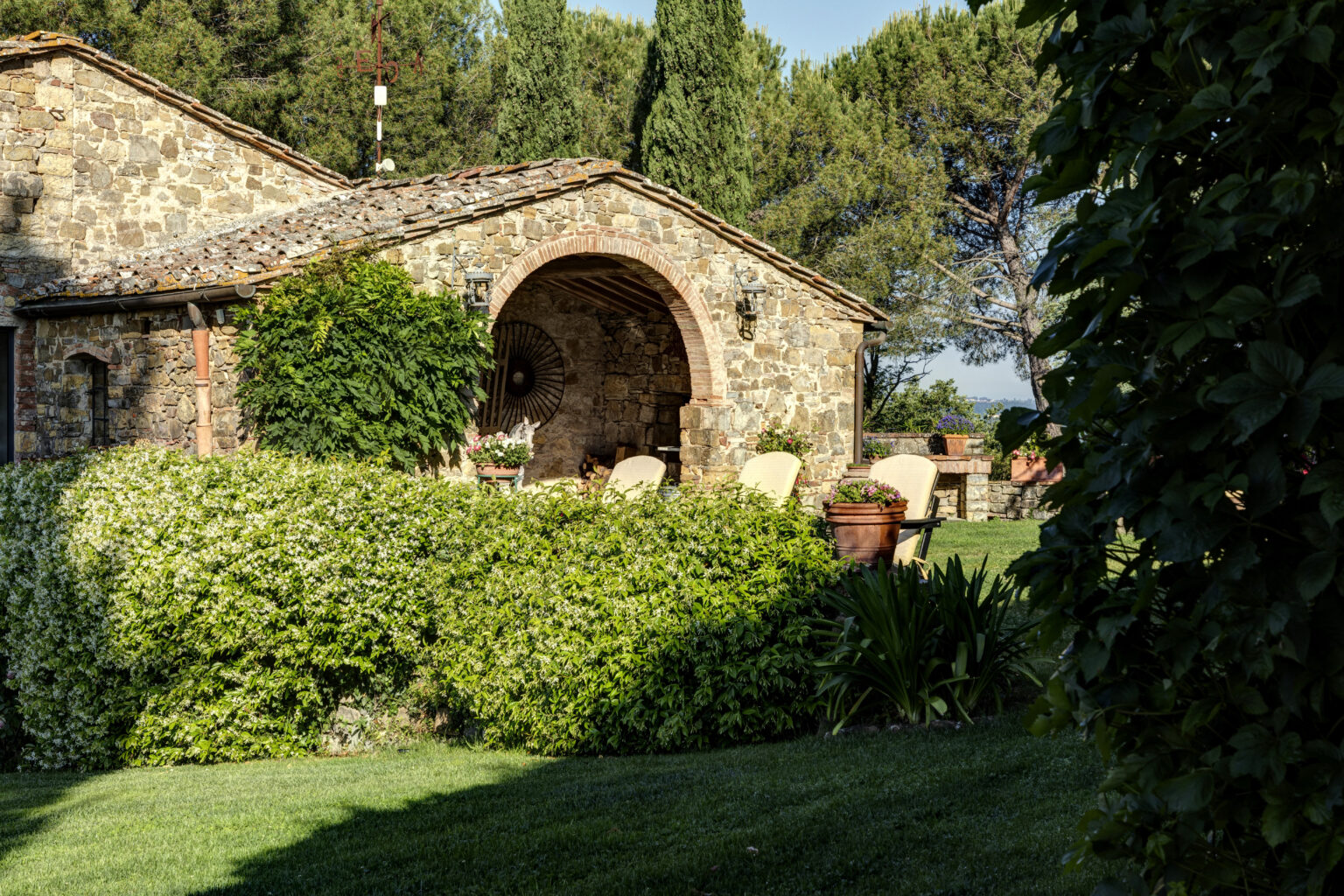 Rustic stone cottage surrounded by lush greenery and flowering shrubs, featuring a cozy patio with chairs and potted plants, set against a backdrop of tall trees in a serene landscape. | Only on SopranoVillas