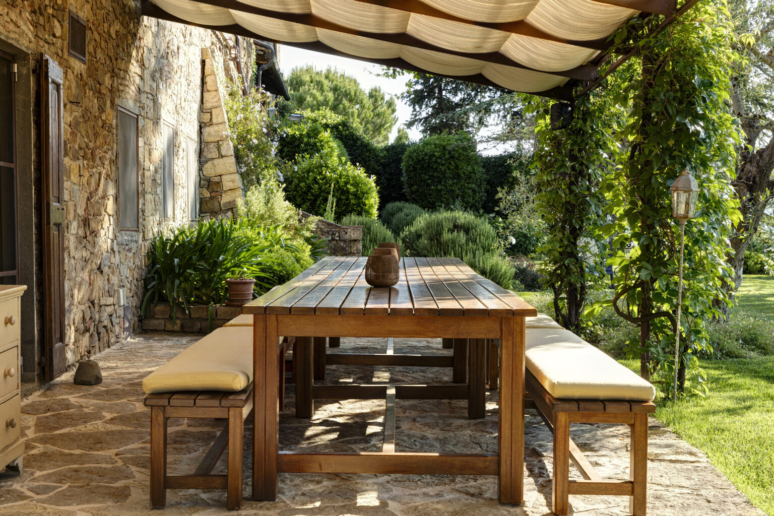Outdoor dining area featuring a rustic wooden table and benches with beige cushions, surrounded by lush greenery and stone walls. The space is shaded by a fabric canopy, creating a serene atmosphere for al fresco meals. | Only on SopranoVillas