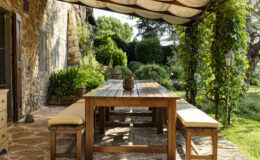 Outdoor dining area featuring a rustic wooden table and benches with beige cushions, surrounded by lush greenery and stone walls. The space is shaded by a fabric canopy, creating a serene atmosphere for al fresco meals.