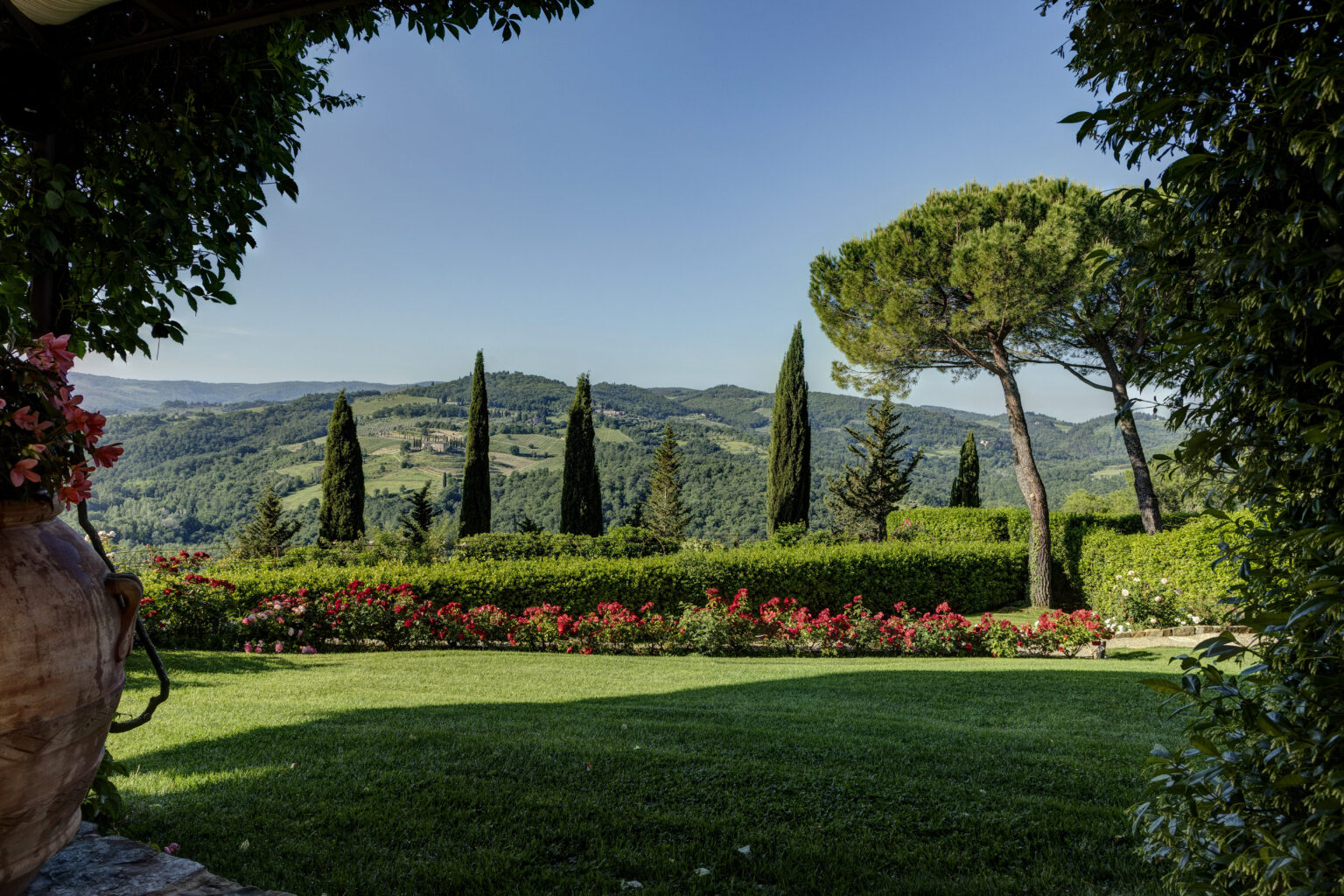 Scenic view of rolling hills in Tuscany featuring tall cypress trees, lush greenery, and vibrant flower beds under a clear blue sky, showcasing the beauty of Italian countryside landscapes. | Only on SopranoVillas