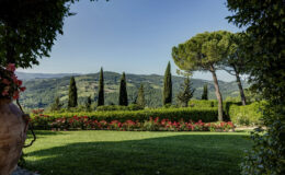 Scenic view of rolling hills in Tuscany featuring tall cypress trees, lush greenery, and vibrant flower beds under a clear blue sky, showcasing the beauty of Italian countryside landscapes.