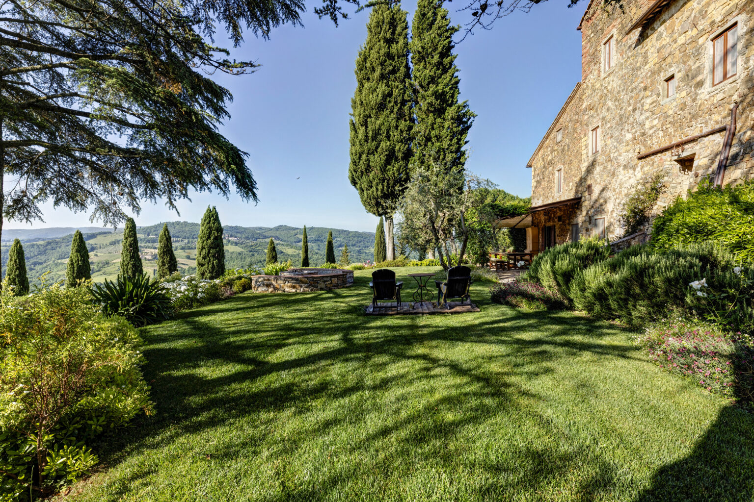 Scenic view of a lush garden with green grass, tall cypress trees, and two black chairs in front of a stone house, overlooking rolling hills under a clear blue sky. Ideal for relaxation and enjoying nature. | Only on SopranoVillas