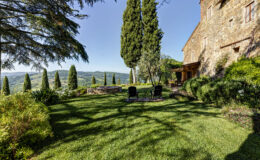 Scenic view of a lush garden with green grass, tall cypress trees, and two black chairs in front of a stone house, overlooking rolling hills under a clear blue sky. Ideal for relaxation and enjoying nature.