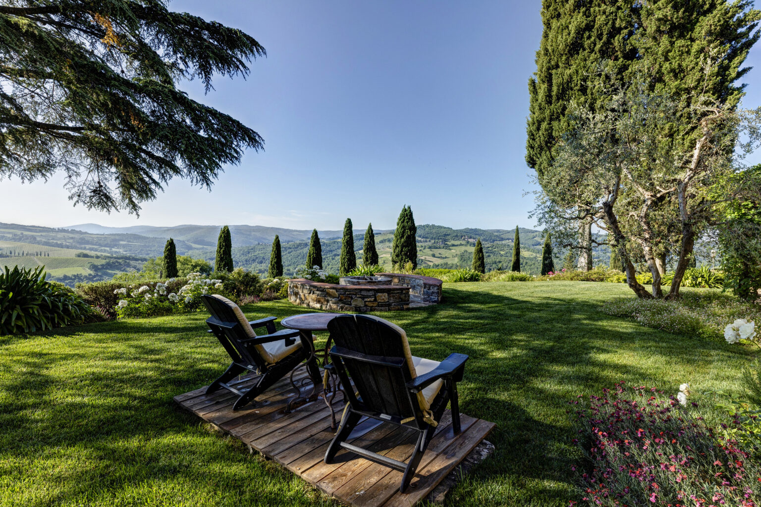 Scenic view of a tranquil garden in Tuscany featuring wooden lounge chairs, a stone fire pit, and manicured greenery with cypress trees and rolling hills in the background under a clear blue sky. | Only on SopranoVillas