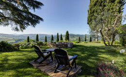 Scenic view of a tranquil garden in Tuscany featuring wooden lounge chairs, a stone fire pit, and manicured greenery with cypress trees and rolling hills in the background under a clear blue sky.