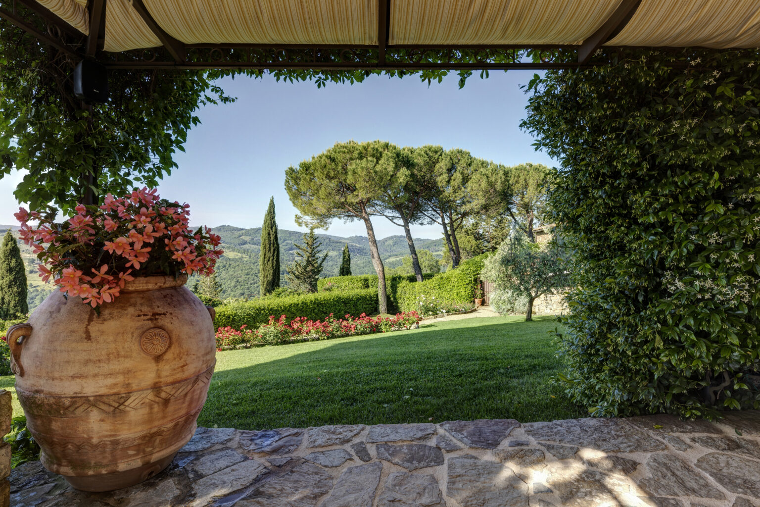 Scenic view from a stone patio featuring a large terracotta pot with pink flowers, lush green lawn, and a backdrop of rolling hills and trees under a clear blue sky. | Only on SopranoVillas
