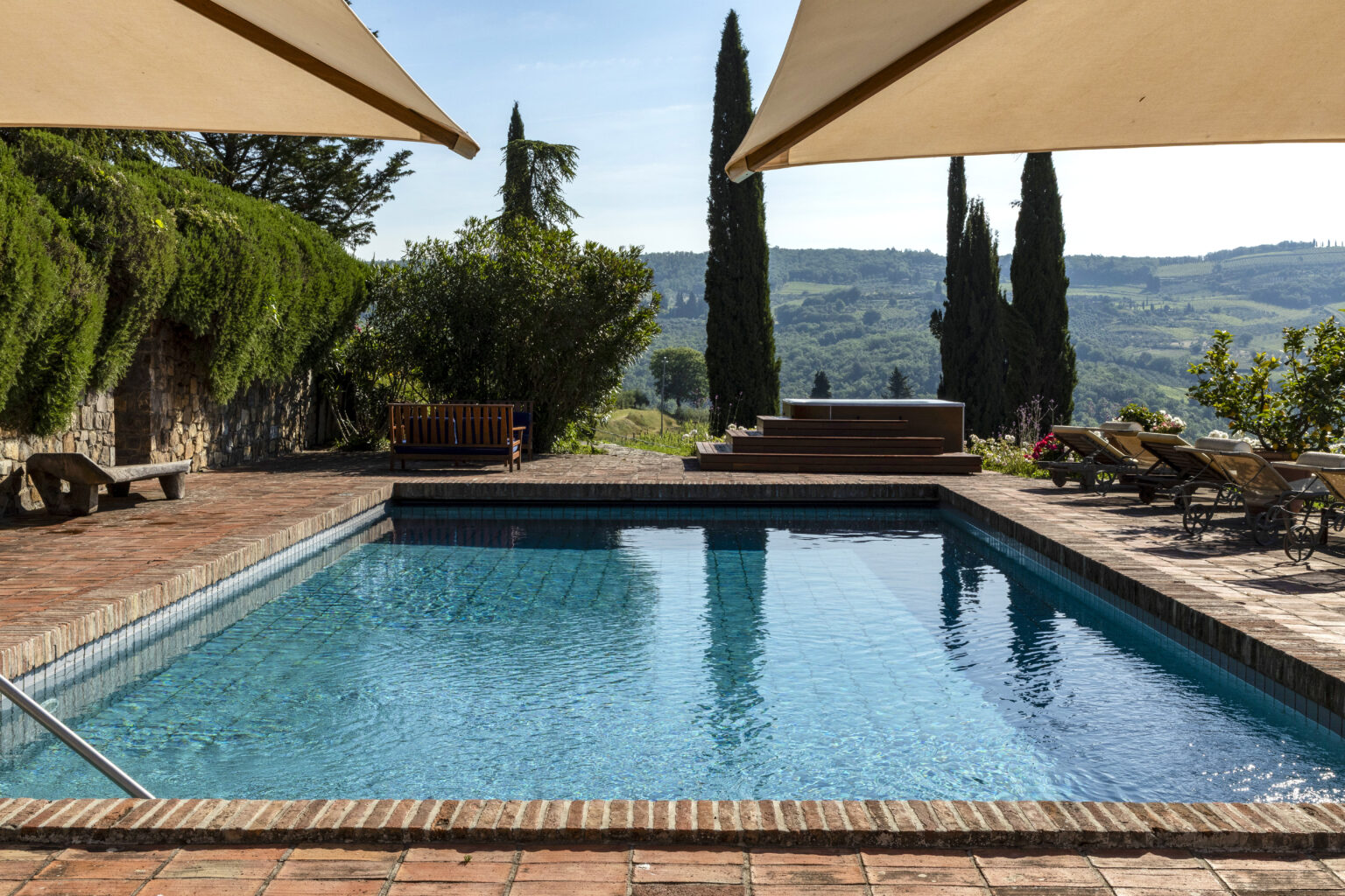 Scenic outdoor swimming pool surrounded by lush greenery and cypress trees, featuring a stone patio with lounge chairs and a wooden bench, set against a picturesque landscape in bright daylight. | Only on SopranoVillas