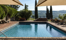 Scenic outdoor swimming pool surrounded by lush greenery and cypress trees, featuring a stone patio with lounge chairs and a wooden bench, set against a picturesque landscape in bright daylight.