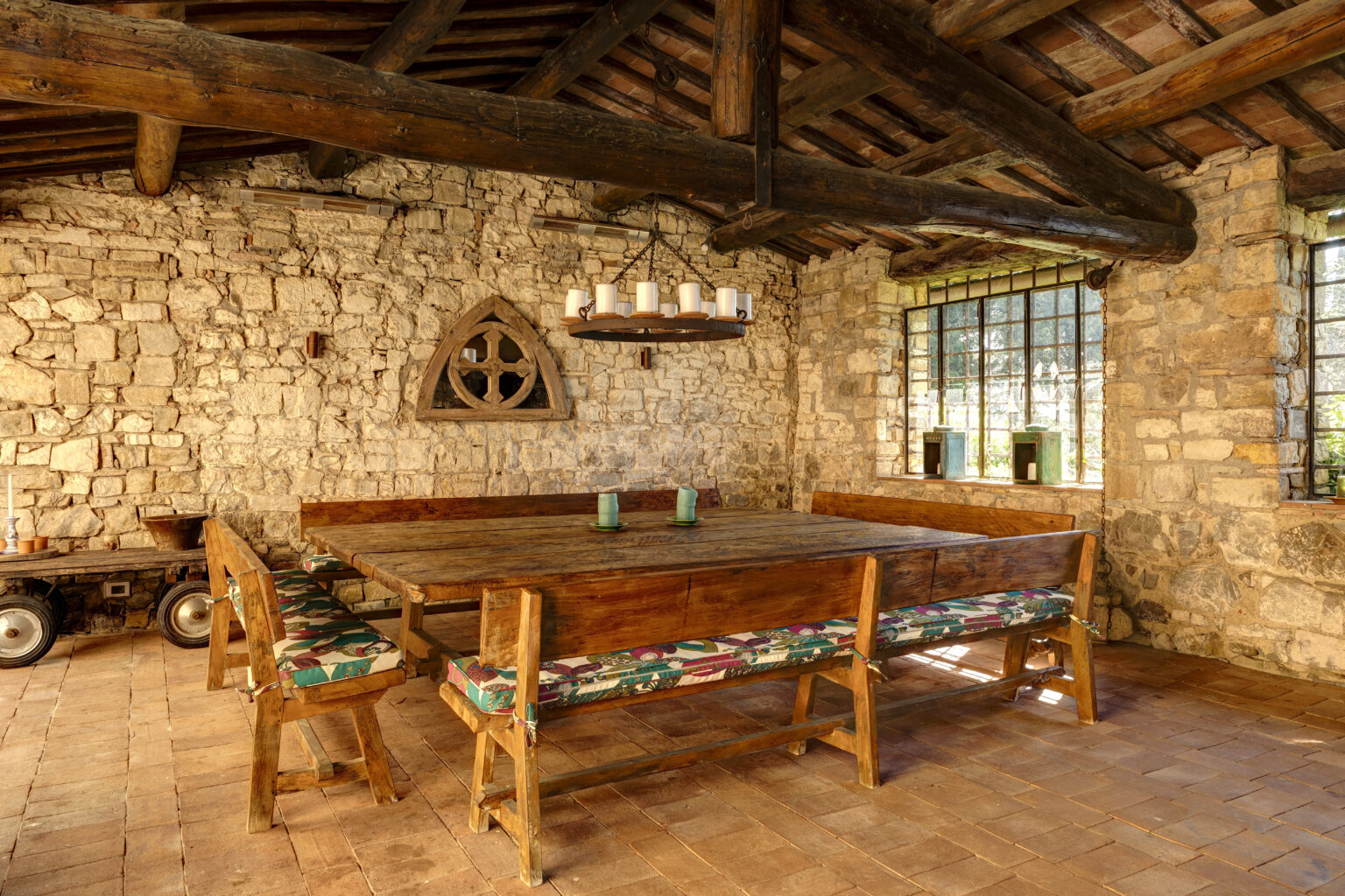 Rustic dining area featuring a large wooden table surrounded by benches with colorful cushions, set against a backdrop of stone walls and exposed wooden beams. Natural light filters through large windows, enhancing the warm, inviting atmosphere of the space. | Only on SopranoVillas