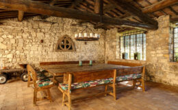 Rustic dining area featuring a large wooden table surrounded by benches with colorful cushions, set against a backdrop of stone walls and exposed wooden beams. Natural light filters through large windows, enhancing the warm, inviting atmosphere of the space.
