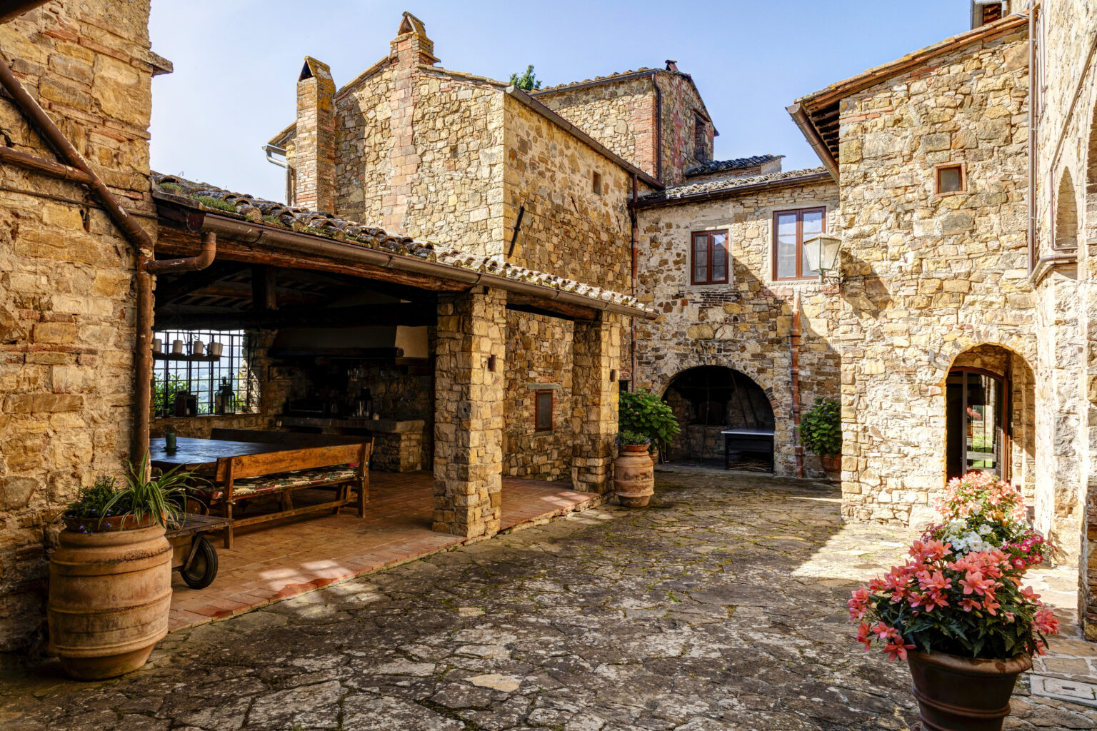 Charming stone courtyard of a rustic villa featuring a wooden dining table, potted plants, and vibrant flowers, bathed in warm sunlight. | Only on SopranoVillas
